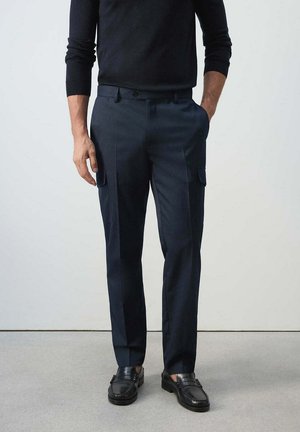 Hombre con pantalones cargo azul marino hechos a medida, camisa negra de manga larga y mocasines de cuero negros, de pie con una mano en el bolsillo.