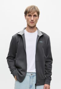 Grå zip-up jakke med grå krage, som har frontglidelås og lommer. Bæreren har på seg en hvit skjorte under og lyseblå jeans.