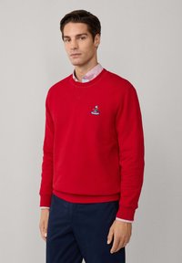 Felpa rossa con collo tondo realizzata in materiale morbido, con un piccolo design ricamato sul petto. Indossata sopra una camicia con colletto.