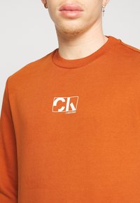 Sudadera de color naranja óxido con cuello acanalado, logo blanco en el pecho y textura de algodón suave. Diseño sencillo con costuras mínimas.