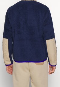 Mörkblå strukturerad tröja med korta ärmar, med en kontrasterande beige panel och lila kant längs nederkanten.