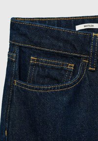 Jeans din denim închis cu cusături contrasting de culoare portocalie, un buzunar frontal și o etichetă pe talie cu inscripția „MATILDA”.