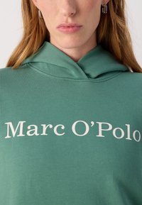 Felpa verde in cotone con tasca a marsupio frontale e logo stilizzato "Marc O’Polo" in crema. Tessuto morbido e vestibilità classica.