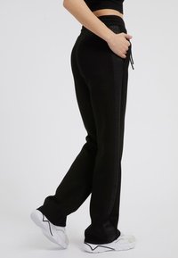 Black sweatpants with a drawstring waistband, side pockets, and a textured stripe running down the sides, paired with white athletic shoes.