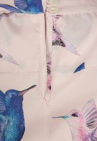 Gros plan sur un tissu rose pâle avec des imprimés de colibris bleus et roses et deux petits boutons blancs près du haut.