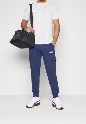 Bílá košile s krátkým rukávem, námořnické cargo kalhoty s kapsami, černá sportovní taška a bílé tenisky s červenými akcenty. Jednoduchý design, ležérní střih.