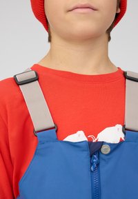 Blauwe overall met grijze bandjes, voorzien van een rits en een zilveren knoop. Draagt men over een rood shirt met een witte grafische print.