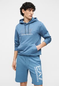 Niebieska bawełniana bluza z kapturem z przednią kieszenią i haftem logo, zestawiona z pasującymi szortami z białym nadrukiem logo po boku.