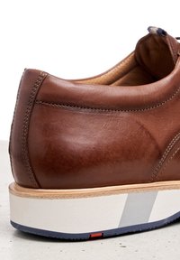 Chaussure en cuir marron avec finition lisse, talon courbé et semelle rayée blanche et marine. Présente des coutures visibles et un design à profil bas.