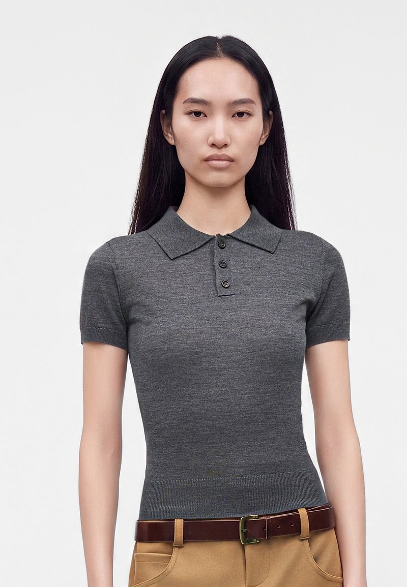 Femme aux longs cheveux foncés porte un polo gris ajusté à manches courtes et un pantalon marron clair avec une ceinture marron foncé.