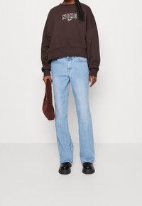 Osoba ubrana w ciemno brązowy sweter Nike, jasnoniebieskie dżinsy, czarne buty, trzyma brązową futrzaną torebkę na tle jednolitego tła.