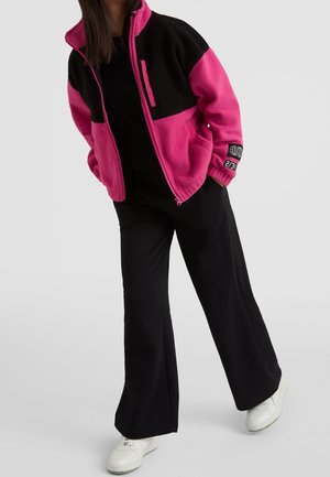 Fleecejakke i pink og sort med lynlås foran og sidelommer, kombineret med sorte bukser med vide ben og hvide sneakers.