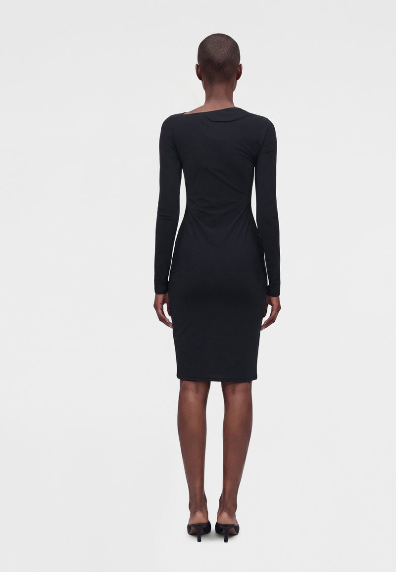Femme debout de dos portant une robe noire ajustée à manches longues et longueur genoux avec des chaussures à talons noires sur un fond uni.