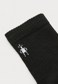 Calcetín negro con textura acanalada y un logotipo de rana de dibujo animado en blanco cerca de la parte superior. El material parece suave y flexible.