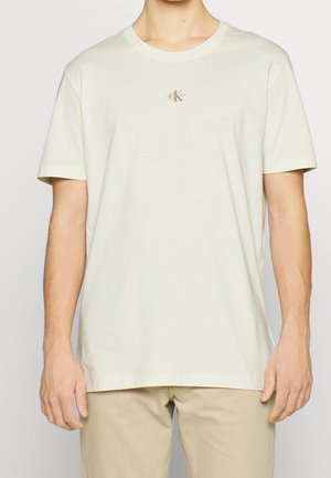 T-shirt en coton beige clair à manches courtes, col rond, avec un petit logo marron sur la poitrine. Coupe régulière, texture lisse.