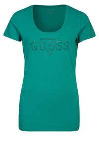 Camiseta de mujer en teal con mangas cortas y escote redondeado. Presenta la palabra "GUESS" en rhinestones en el pecho. Material de algodón suave.