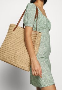 Sac fourre-tout tissé beige avec motif texturé et anses en cuir marron, porté par une personne vêtue d'une robe verte à fleurs.