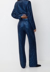 Sukienka z cekinami w ciemnoniebieskim kolorze, składająca się z bluzki z długim rękawem i spodni z szerokimi nogawkami. Materiał jest błyszczący, z teksturowaną, odbijającą światło powierzchnią.