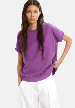 Fioletowy bawełniany t-shirt z krótkimi rękawami, okrągłym dekoltem i luźnym krojem, w zestawieniu z białymi spodniami w zakładki. Gładka tekstura, casualowy design.