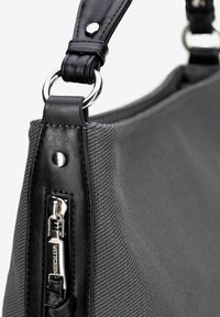 Šedá látková kabelka s černými koženými akcenty, obsahující zipovou kapsu, kovové detaily a texturovaný povrch. Součástí je nastavitelné rameno.