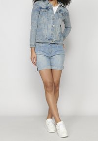 Nainen, joka käyttää vaaleansinistä kukkakirjailtua farkkutakkia ja siihen sopivia ylös käärittyjä shortseja valkoisten tennariden kanssa, seisoo yksinkertaista taustaa vasten.