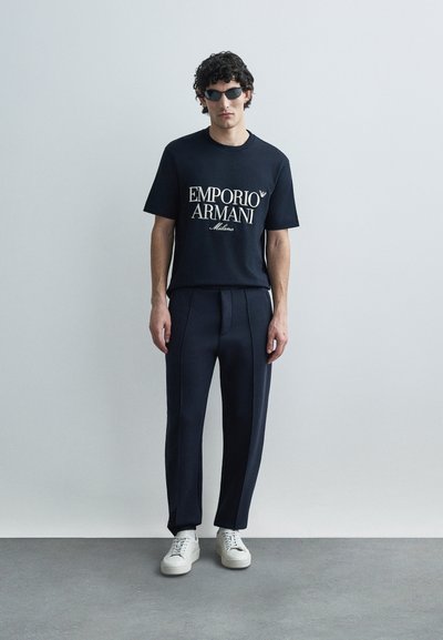 Μαύρο βαμβακερό t-shirt με λευκό γράφημα "EMPORIO ARMANI", συνδυασμένο με σκούρο, κομψό παντελόνι και λευκά αθλητικά παπούτσια, με μοντέρνο σχεδιασμό.