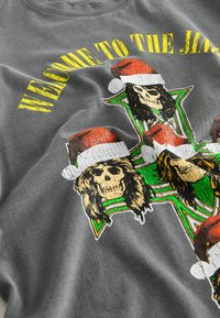 Szürke póló, amelyen grafika látható: koponyák karácsonyi sapkával, zöld és sárga díszítéssel, valamint kopottas felirattal, amely így szól: "ÜDV A JINGLE-BEN."