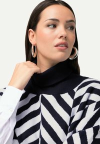 Maglione a righe blu navy e bianche con collo alto, trama morbida e vestibilità comoda. Il modello indossa grandi orecchini a cerchio argentati e una camicia bianca.