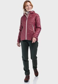 Bordeauxrote isolierte Jacke mit Kapuze, ausgestattet mit einem diagonalen Steppmuster und einem weißen Reißverschluss, kombiniert mit grünen Hosen und braunen Wanderschuhen.