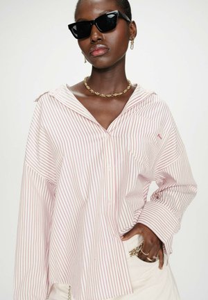 Personne portant une chemise oversize rayée rose et blanche, des lunettes de soleil noires, un collier en chaîne dorée, des boucles d'oreilles et des bagues, avec la main dans la poche.