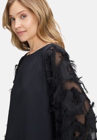 Top nero con maniche trasparenti caratterizzato da motivi a frange nere sovrapposte. Design con scollatura rotonda semplice, con una texture di tessuto liscia sul corpo.