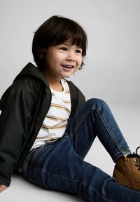 Un copil într-o jachetă verde închis, purtând o cămașă albă cu dungi, blugi și cizme maro, așezat pe un fundal neutru.