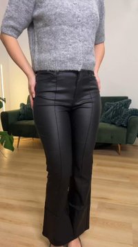 Calças pretas de couro sintético com corte flare, apresentando costuras contrastantes e textura suave. Usadas com uma camisola cinzenta de malha de manga curta.