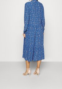 Blå blommig klänning med långa ärmar, samlad midja och lager-på-lager kjol. Stylar med ljusrosa klacksandaler. Slät tygtextur.