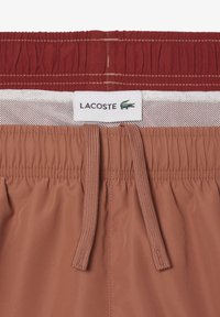 Bañador con cinturilla elástica en color óxido, que cuenta con un cordón ajustable, forro de malla y una prominente etiqueta del logo de Lacoste. Textura suave.