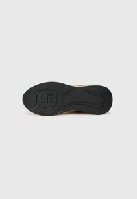 Suola in gomma nera con un motivo a zigzag e logo. La parte superiore è beige, caratterizzata da un design senza cuciture e liscio, senza chiusure visibili.
