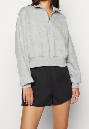 Femme portant un sweat-shirt gris clair à demi-fermeture éclair et un short de sport noir sur fond blanc uni.