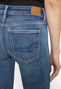 Vaqueros de mezclilla azul oscuro con un diseño ajustado, que cuentan con un bolsillo trasero con detalles de costura y una etiqueta de cuero marrón con la inscripción "American Eagle."