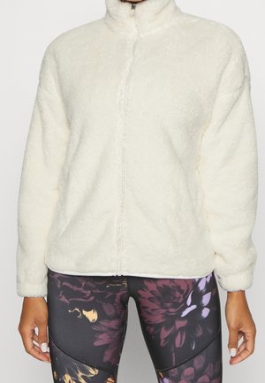 Personne portant une veste sherpa blanche zippée et des leggings noirs avec de grands motifs floraux violets et dorés.
