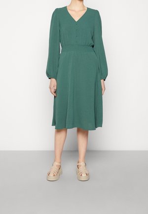 Femme portant une robe verte mi-longue avec des manches longues bouffantes, un col en V boutonné, une taille cintrée et des sandales à plateforme beiges.