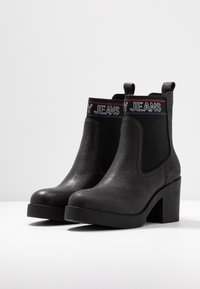 Czarne botki na obcasie z elastycznymi panelami bocznymi, teksturowana sztuczna skóra oraz kontrastującą wstążką z logo z napisem "MY JEANS" w białym i czerwonym kolorze.