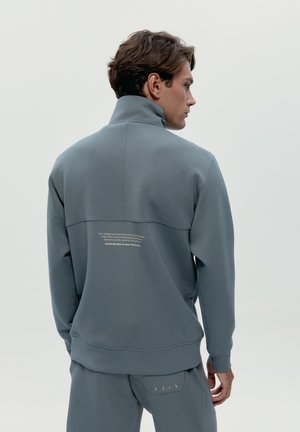 Joven de espaldas vistiendo sudadera y pantalones azul grisáceo a juego con un pequeño texto blanco en la parte superior de la espalda y el bolsillo.