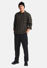 Chemise à carreaux verte et noire avec boutons, poche avant ; associée à un pantalon cargo noir et des baskets noires. Portée avec un bonnet noir.