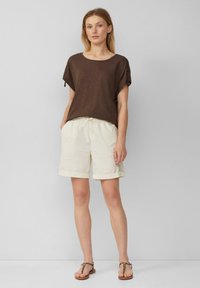 Bruine, korte mouwen top met een brede halslijn, gecombineerd met witte shorts met een elastische taille en opgerolde boorden. Gestrikte sandalen maken de look compleet.