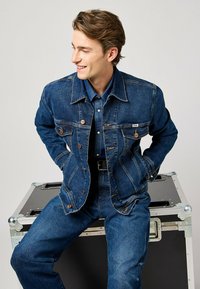 Tummansininen farkkutakki perinteisellä leikkuulla, jossa on metallinuppinepparit, rintataskut ja kontrastiväriset ompeleet. Päällä farkkupaita.