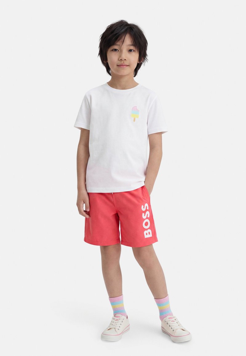 Παιδί που στέκεται, φορώντας λευκό μπλουζάκι με γραφική εικόνα παγωτού, κόκκινα σορτς με τη λέξη "BOSS", ριγέ παστέλ κάλτσες και λευκά αθλητικά παπούτσια.