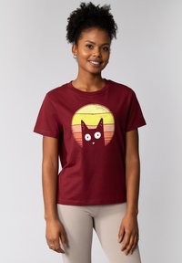 Bordeauxrotes Baumwoll-T-Shirt mit einem Grafikdesign eines schwarzen Katers mit großen Augen vor einem gelb-orangefarbigen, kreisförmigen Sonnenuntergang. Kurze Ärmel.