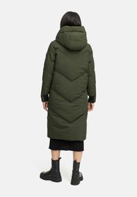 Abrigo largo acolchado verde con capucha, que presenta un patrón en forma de chevrón, y una falda de canalé negra y botas negras gruesas visibles en la parte inferior.