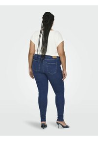 Jeans ajustados en denim azul oscuro con cintura alta, que cuentan con dos bolsillos traseros y una etiqueta de cuero en la cinturilla. Combinados con una blusa blanca.