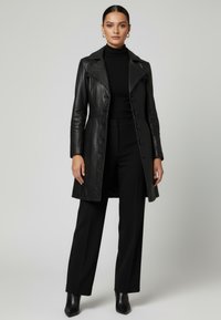 Kvinna står klädd i en svart läderknäppt kappa, svart polotröja, vida byxor, ankelboots och guldfärgade ringörhängen mot en enfärgad bakgrund.